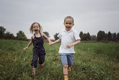 Portrait of happy wet siblings holding hands while running on grassy field against sky at park during rainy season