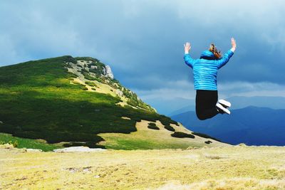 Rear view full length of woman with arms raised jumping over field against cloudy sky
