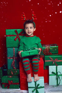 Baby boy in a green t shirt and with deer antlers sits with gifts on a red background