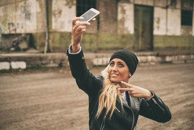 Fashionable young woman wearing leather jacket taking selfie with smart phone