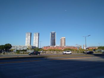 Cars on road by buildings against clear blue sky