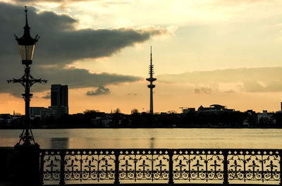 Silhouette heinrich-hertz-turm and cityscape by river against sky during sunset