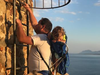 Side view of father climbing ladder on wall while carrying son against sky