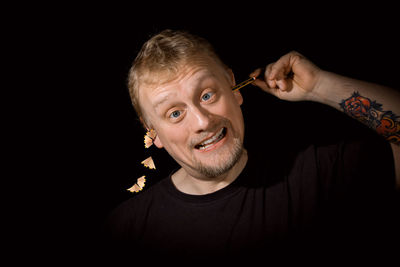 Portrait of smiling man inserting pencil in ear against black background