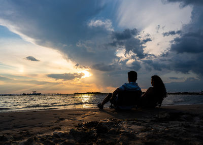 Couple sitting on beach against sky during sunset