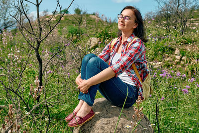 Happy carefree woman sitting on big stone in a spring meadow with beautiful field flowers. 