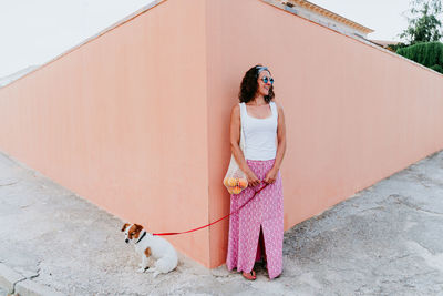 Full length of woman with dog standing on footpath against wall