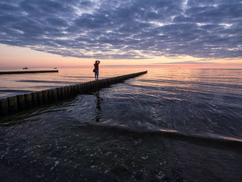 Man standing on stepping stone at beach against sky during sunset