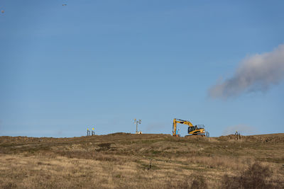 A yellow excavator on the top of the hill with a blue sky and a gray cloud behind