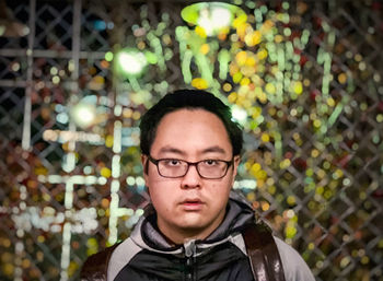 Portrait of a man wearing eyeglasses against metal fence, street and neon lights.