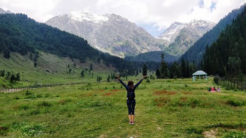 Rear view of woman with arms raised looking at mountains while standing on grassy land