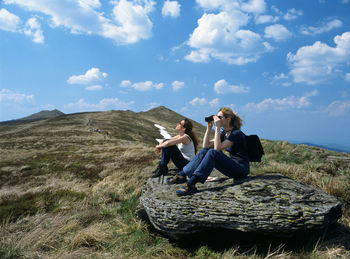 Young man using mobile phone while sitting on mountain against sky