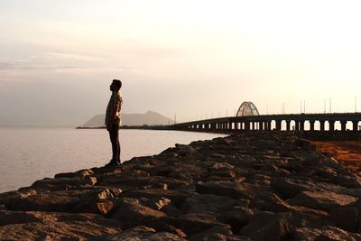 Man standing on rock by sea against sky during sunset