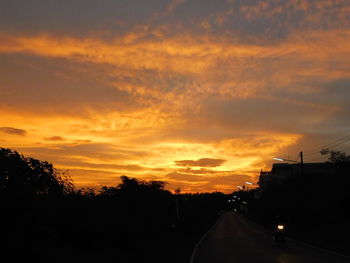 Road amidst silhouette trees against dramatic sky during sunset