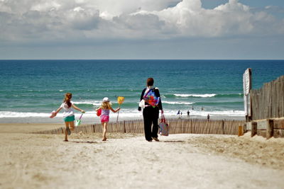 Rear view of woman with daughters walking at beach against cloudy sky