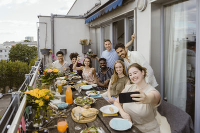 Smiling woman taking selfie with group of friends through smart phone while celebrating dinner party in balcony