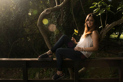 Full length of smiling woman sitting on railing by trees