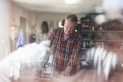 Senior man using circular saw in workshop seen through glass window