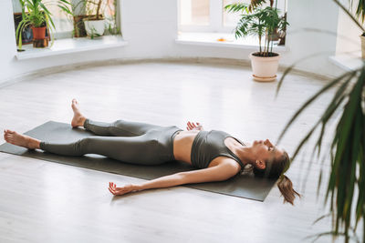 Young fit woman practice yoga doing savasana in light yoga studio with green house plant