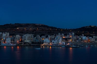 Illuminated buildings by sea against clear blue sky at night