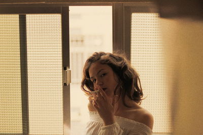 Portrait of young woman smoking while standing against window at home