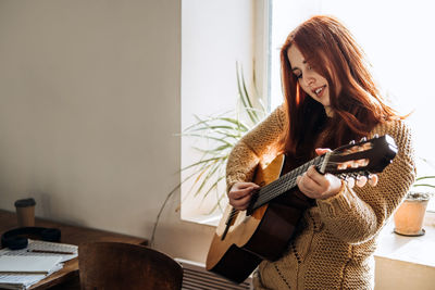 Indoor cozy hobbies for winter, autumn cold season. redhead woman playing acoustic guitar and