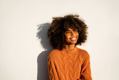 Full length of smiling ethnic woman with curly afro hair wearing vivid orange sweater looking away leaning against white modern concrete wall background