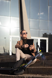 Vertical view of young bussinessman reading an ebook in the stair with an electric scooter