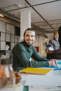 Smiling young male student sitting with books and files at table in community college