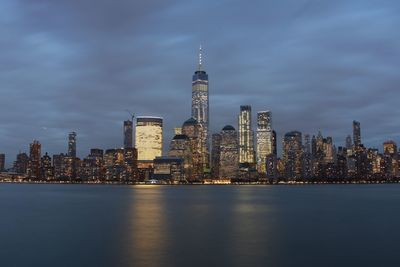 Illuminated city at waterfront against cloudy sky