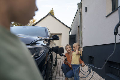 Woman holding electric car charger plug looking at daughter in front yard