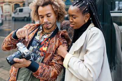 Young stylish african american male photographer with photo camera showing pictures to female model during session on city street