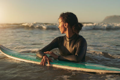 Young woman with surfboard looking away in sea during sunset