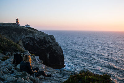 Man sitting on rock by sea against sky during sunset