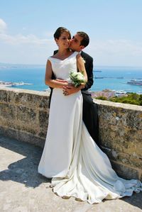 Groom kissing bride by retaining wall against sea