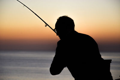 Silhouette man fishing at beach against sky during sunset