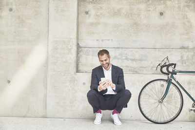 Smiling businesssman with bicycle at concrete wall looking at cell phone