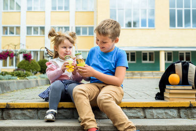 Full length of girl and boy holding drink sitting on steps against school building