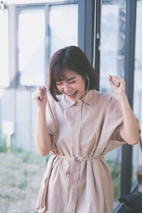 Smiling young woman listening music while standing by window at home