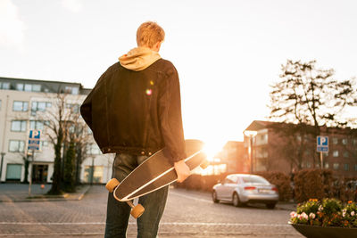 Rear view of young man holding skateboard while standing on sidewalk in city