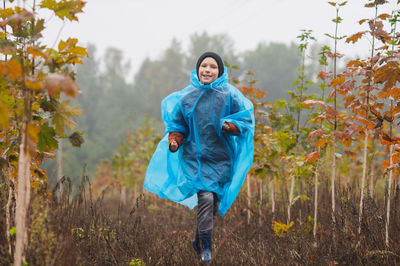 A boy runs in a blue raincoat near small maple trees in autumn
