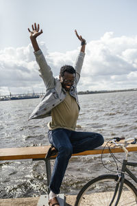 Young man with bicycle, sitting on railng by the sea, pretending to fly