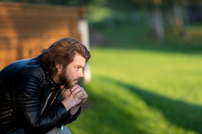 Pensive serious young bearded man wear black leather jacket sitting and thinking outdoors