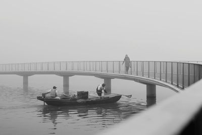 People fishing in boat on lake while woman walking on footbridge against sky