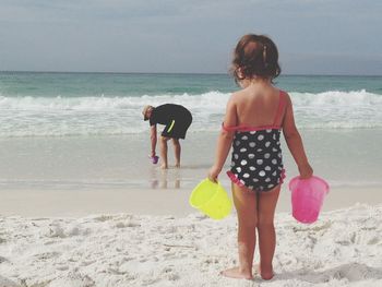 Rear view of child standing thinking on beach with sand pails watching adult search for seashells