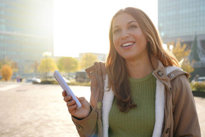 Woman holding mobile phone in the city smiling looking to the side and staring away thinking