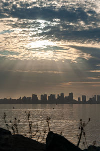 Silhouette buildings by lake against cloudy sky during sunset