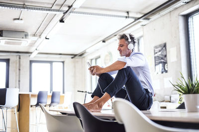 Mature businessman with smartphone and headphones sitting barefoot on desk in office barefoot