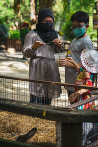 Children wearing face masks are feeding the rabbit at the wooden cage.