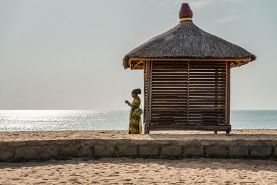 African woman standing next to a bungalow alone wearing african clothes on the coast of africa